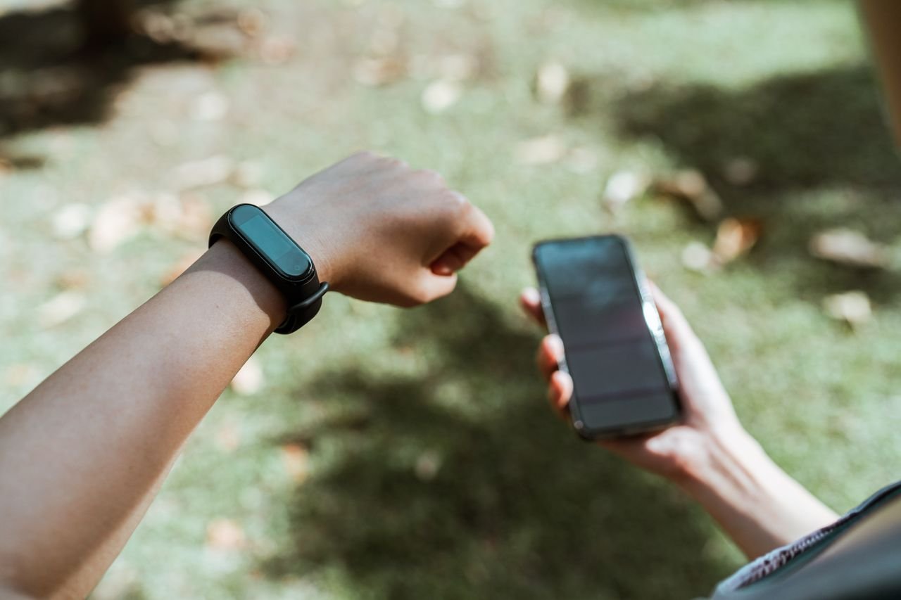 Top view of crop anonymous person demonstrating modern plastic bracelet with small display while surfing internet on cellphone sitting in tranquil park covered with faded leaves in daylight